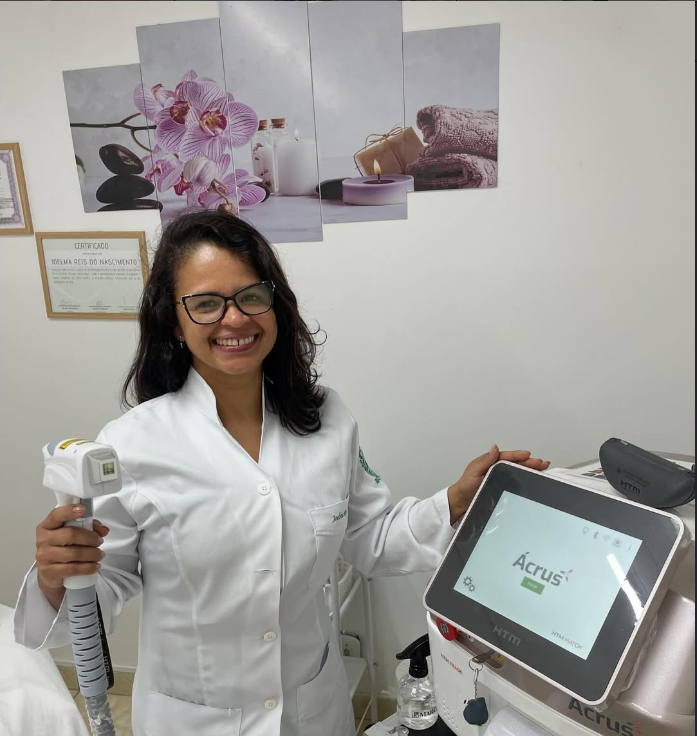 Joelma sorrindo, de jaleco branco, segurando a caneta de um equipamento de laser em sua sala de atendimento