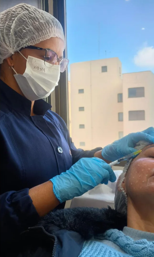 Joelma, de touca, máscara e luvas, realizando um procedimento facial em uma cliente